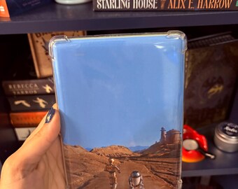 Puede incluir: Una funda transparente para teléfono con un paisaje desértico con dos figuras y un droide. La parte superior de la funda tiene un diseño de cielo azul. La funda se sostiene frente a una estantería con libros y una vela.