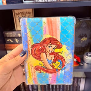 May include: Clear protective case featuring a colorful design with a mermaid and a yellow fish. The case has a blue scale pattern and a rainbow background. The mermaid has red hair and is wearing a blue top.