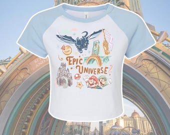 Puede incluir: Top corto blanco y azul claro con un diseño gráfico que presenta un dragón, una varita mágica, un castillo y el texto "Epic Universe?" La camiseta también incluye imágenes de Mario y Luigi.
