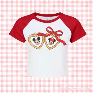 May include: White and red baseball tee shirt with a graphic of Mickey and Minnie Mouse inside heart-shaped lockets connected by a red ribbon with a bow.