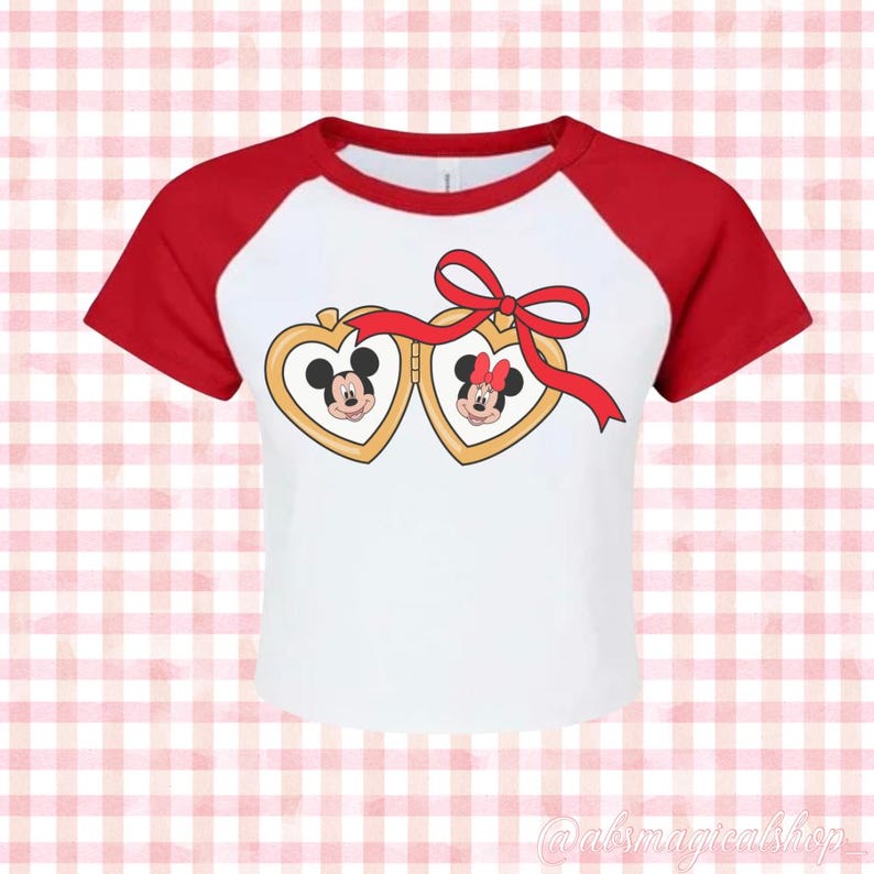 May include: White and red baseball tee shirt with a graphic of Mickey and Minnie Mouse inside heart-shaped lockets connected by a red ribbon with a bow.
