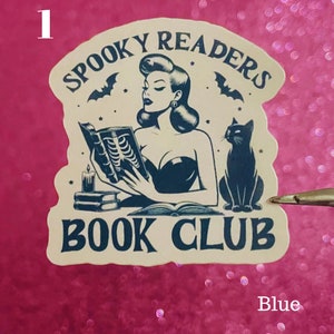 May include: A beige sticker with a black and white illustration of a woman reading a book with a black cat sitting beside her. The sticker says "Spooky Readers Book Club" in a black font.