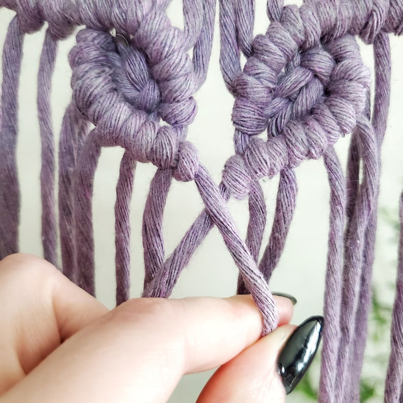 May include: Close-up of a hand tying a knot in a purple macrame wall hanging. The knot is being tied in the center of the image, with the strands of yarn extending out to the sides.