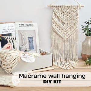 May include: A macrame DIY kit featuring a white cardboard box containing a bundle of off-white cotton cord, a knot guide, and a pattern booklet labelled CORD + QUARTZ. A finished off-white macrame wall hanging with a chevron pattern hangs nearby.