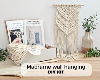 Puede incluir: Un kit de manualidades de macramé que incluye una caja de cartón blanca con un ovillo de cuerda de algodón color crema, una guía de nudos y un folleto de patrones con la etiqueta CORD + QUARTZ. Un tapiz de macramé color crema cuelga cerca.