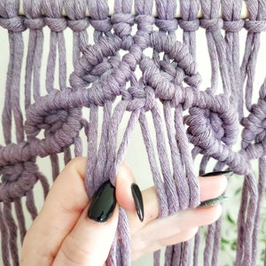 May include: Close-up of a macrame wall hanging with a purple cord. The cord is tied in a knot pattern, creating a series of small circles. A hand is holding the cord, showing the detail of the knot.