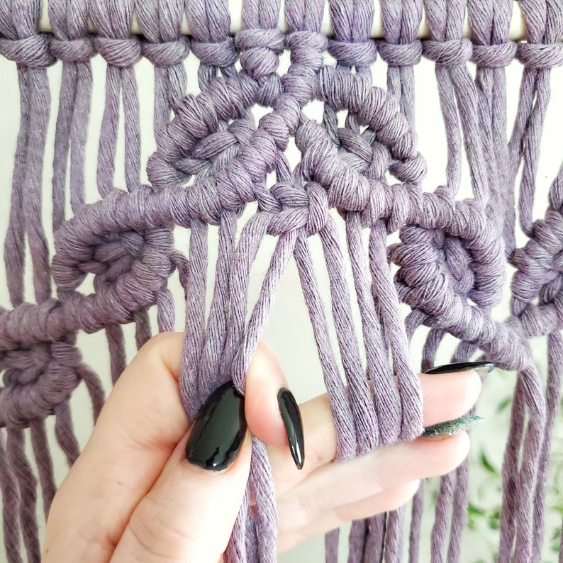 May include: Close-up of a macrame wall hanging with a purple cord. The cord is tied in a knot pattern, creating a series of small circles. A hand is holding the cord, showing the detail of the knot.