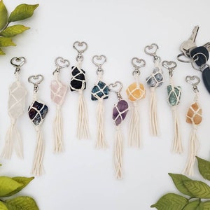 May include: A collection of ten crystal keychain charms, each featuring a different crystal wrapped in macrame and attached to a silver heart-shaped key ring. The crystals include clear quartz, amethyst, rose quartz, black obsidian, green aventurine, citrine, and blue apatite.