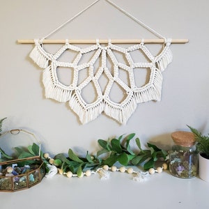 May include: A white macrame wall hanging with a geometric design and fringe. The hanging is attached to a wooden dowel rod.