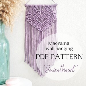 May include: Purple macrame wall hanging with a heart-shaped design. The hanging is labeled "Macrame wall hanging PDF PATTERN 'Sweetheart'".