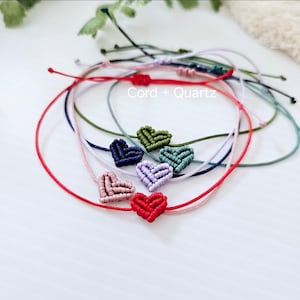 May include: A set of seven colourful friendship bracelets with knotted heart charms. The bracelets are made with different coloured cords and feature a variety of heart colours, including red, green, blue, and purple. The text "Cord + Quartz" is visible in the image.