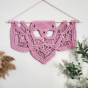 May include: A pink macrame wall hanging with a geometric design. The hanging is made with thick cotton cord and is attached to a wooden dowel.