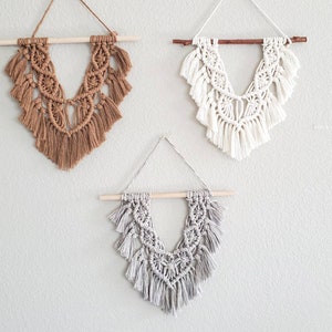 May include: Three macrame wall hangings with a V-shaped design. The hangings are made with natural fibers and have a fringe detail. The colors are brown, white, and gray.