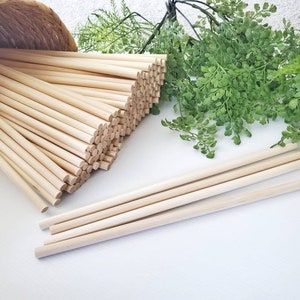 May include: A bunch of natural wooden dowels, approximately 12 inches long, are stacked in a woven basket. A few dowels are laying on a white surface.