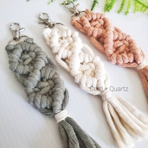 May include: Three macrame keychains with tassels in green, white, and peach colors. The keychains are made with a knotting technique and have silver key rings. The text "Cord + Quartz" is visible in the image.