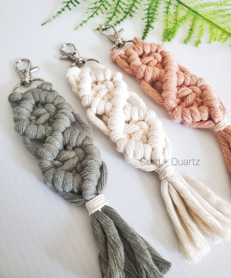 May include: Three macrame keychains with tassels in green, white, and peach colors. The keychains are made with a knotting technique and have silver key rings. The text "Cord + Quartz" is visible in the image.