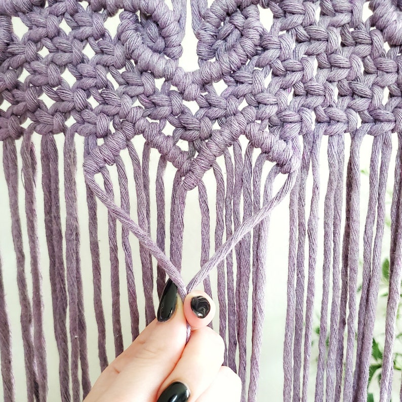 May include: A close-up of a purple macrame wall hanging with a heart shape in the center. The macrame is made with a thick cord and has a fringe at the bottom.