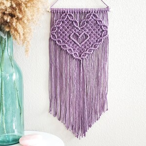 May include: A purple macrame wall hanging with a heart design. The hanging has a fringe of tassels at the bottom.