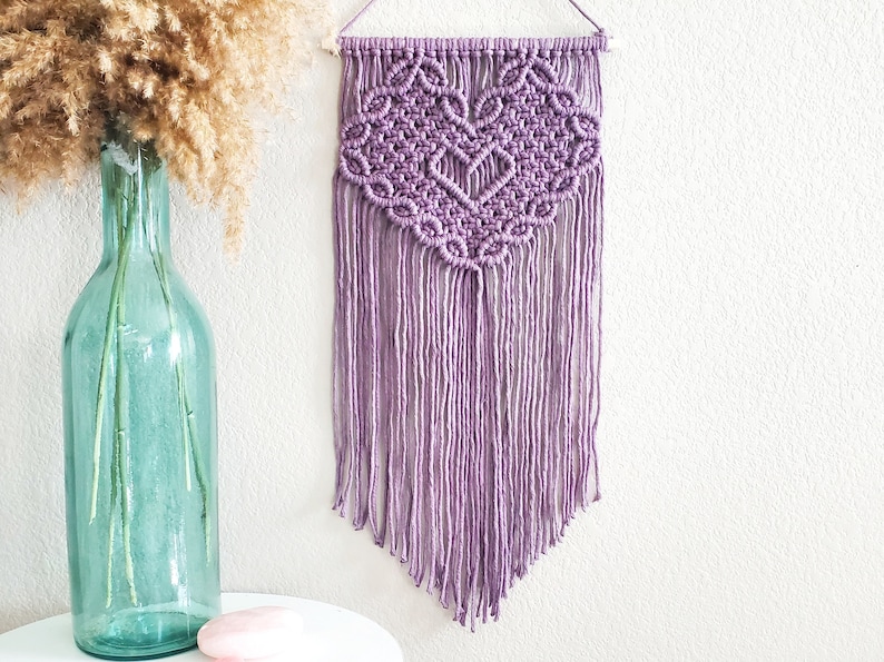May include: A purple macrame wall hanging with a heart design. The hanging has a fringe of tassels at the bottom.