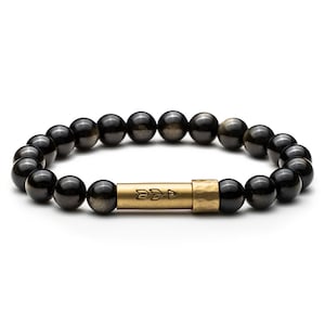 Peut inclure: Un bracelet élastique composé de perles en obsidienne noire polie avec un éclat métallique. La pièce centrale est un tube cylindrique en laiton présentant une texture martelée à une extrémité et une flèche gravée sur sa surface lisse.