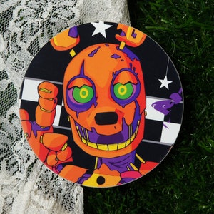 May include: Round sticker featuring a cartoon character with orange skin, purple and green eyes, and a toothy grin. The character is set against a black and white checkered background with stars.