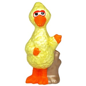 Puede incluir: Figura amarilla de un pájaro de dibujos animados con pies, pico y puntas de las alas naranjas. El pájaro tiene marcas rojas en los ojos y está de pie sobre una base marrón. La figura es una representación caprichosa de un personaje infantil popular.