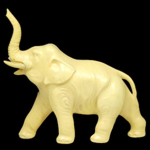 May include: Ivory-colored elephant figurine with trunk raised and tusks visible. The sculpture has detailed carved patterns on its body and is set against a black background. A decorative piece.