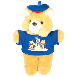 May include: A plush teddy bear ornament with a blue hat and sweater. The sweater features a snowman and two bears. The bear is tan with a white muzzle and black eyes. A blue loop is attached to the hat for hanging.