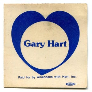 May include: A vintage campaign button featuring a blue heart shape with the name "Gary Hart" in the center. The text "Paid for by Americans with Hart, Inc." is printed below the heart. The button has a worn, aged appearance.