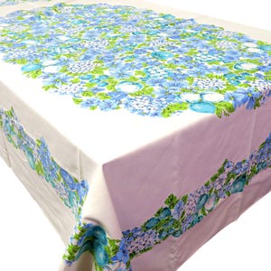 May include: A white rectangular tablecloth with a floral pattern. The pattern features blue and turquoise flowers with green leaves. The flowers are clustered in the center and along the edges of the tablecloth.