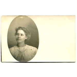 Puede incluir: Una fotografía vintage en tonos sepia impresa en una tarjeta rectangular. La imagen presenta un retrato ovalado de una persona con cabello ondulado, vistiendo una blusa clara con detalles de encaje intrincados alrededor del cuello y el pecho.