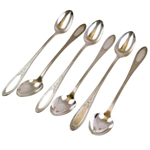 May include: Six silver spoons with elongated handles and oval bowls are arranged diagonally against a white background. The spoons have decorative detailing on the handles, with some featuring openwork designs. The spoons are reflective, showing the surrounding environment.