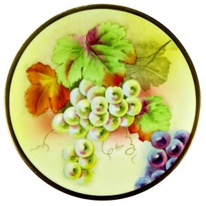 Puede incluir: Un plato decorativo redondo con borde dorado. El plato presenta un diseño pintado a mano de uvas verdes y moradas, hojas verdes y naranjas y delicadas vides sobre un fondo amarillo.