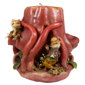 May include: A carved candle in shades of red and brown, featuring a whimsical design. The candle depicts a tree trunk with a small figurine of a man and a cat. Decorative floral accents are also present.