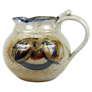May include: A speckled beige ceramic pitcher with a rounded body, a curved handle, and a pouring spout. The surface features a decorative abstract pattern in shades of blue, brown, and tan, with a dark blue rim accenting the top edge of the vessel.