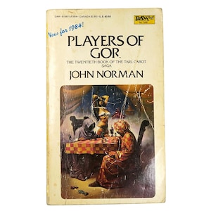 May include: A vintage paperback book titled "Players of Gor" by John Norman. The cover art features a fantasy scene with figures around a checkered table. The book is yellowed with age and has the text "New for 1984!" and the publisher's logo.