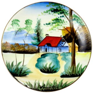 May include: A round decorative plate with a hand-painted landscape scene. The image features a white house with a red roof, trees, and a body of water. The plate has a gold rim and a signature at the bottom.