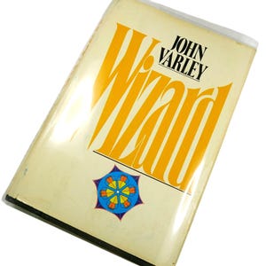 May include: A vintage paperback book titled "Wizard" by John Varley. The cover features large, yellow stylized text and a colorful geometric design. The book has a cream-colored background and is encased in a clear plastic cover.