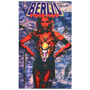 May include: A rectangular graphic print featuring the text BERLIN FRIDAYS in bold blue and red lettering. The central figure is a person in a red-toned outfit with a skull emblem on the chest, set against a textured, shimmering purple background.