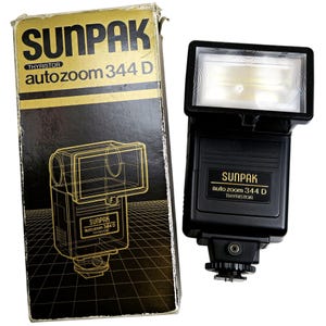 Puede incluir: Un flash Sunpak auto zoom 344 D vintage y su caja original. El flash es negro con un cabezal rectangular y el texto "SUNPAK auto zoom 344 D THYRISTOR". La caja es negra y dorada con el texto "SUNPAK auto zoom 344 D THYRISTOR".