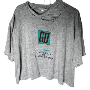 May include: A heather gray short-sleeve t-shirt with a distressed, ripped neckline. The front features a graphic with the word GO in red letters inside a teal square, followed by the text UNIVERSITY OF RHODE ISLAND in dark blue lettering.