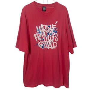 May include: A red t-shirt featuring a white and blue paint splatter design and the text "MARIE FRANCOIS GREAP" in white. The shirt has short sleeves and a crew neck.