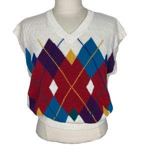 May include: A sleeveless, white V-neck sweater vest with a colorful argyle pattern. The argyle design features red, blue, purple, yellow, and green diamonds. The vest is displayed on a mannequin.