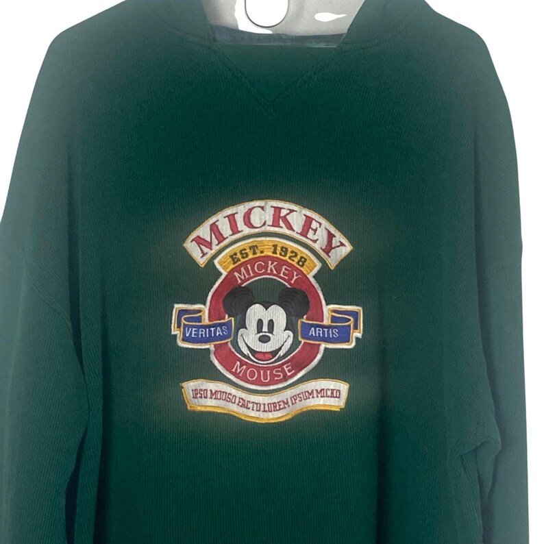 May include: A dark green hooded sweatshirt featuring an embroidered Mickey Mouse graphic on the chest. The design includes the text MICKEY EST. 1928, VERITAS ARTIS, and MOUSE, along with Latin-style text at the bottom of the circular emblem.