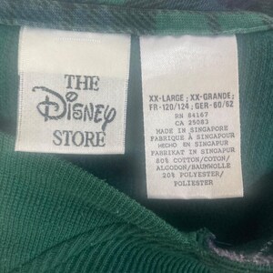 May include: A dark green garment featuring two white fabric tags. The left tag displays The Disney Store logo. The right tag lists size XX-Large, RN 84167, CA 25083, Made in Singapore, and a material composition of 80% cotton and 20% polyester.