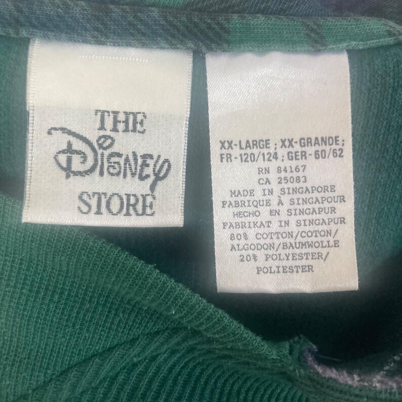 May include: A dark green garment featuring two white fabric tags. The left tag displays The Disney Store logo. The right tag lists size XX-Large, RN 84167, CA 25083, Made in Singapore, and a material composition of 80% cotton and 20% polyester.