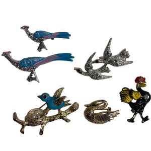 May include: A set of six vintage bird brooches. The brooches are made of metal and feature a variety of bird designs, including a peacock, a swan, a robin, and a rooster. The brooches are decorated with rhinestones and enamel.