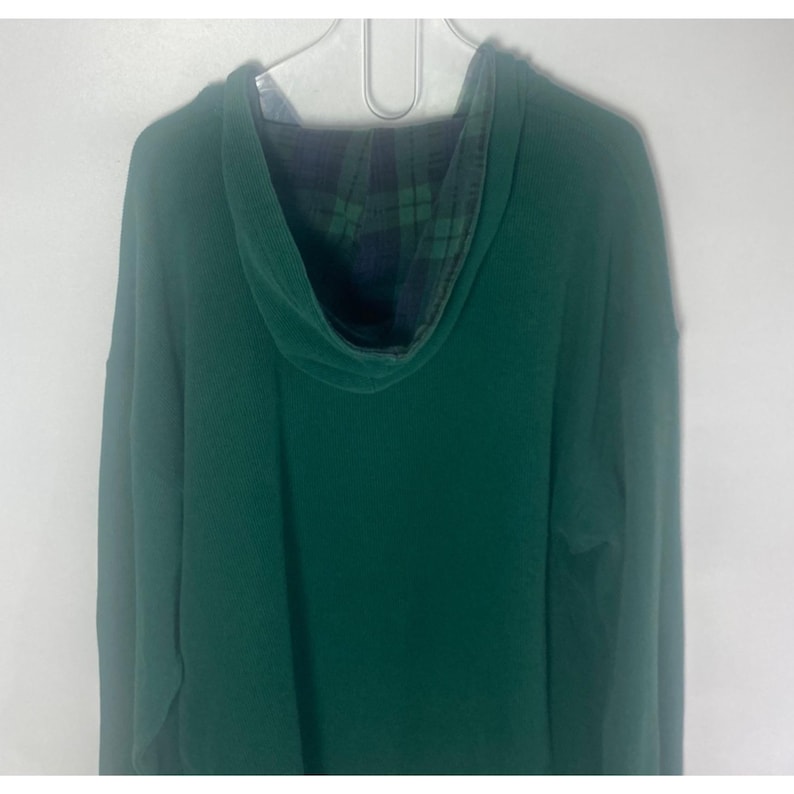 May include: A solid dark green long-sleeve hooded sweatshirt featuring a hood lined with a dark green and navy blue plaid pattern. The garment is displayed on a white hanger and features a standard hood construction with long sleeves.