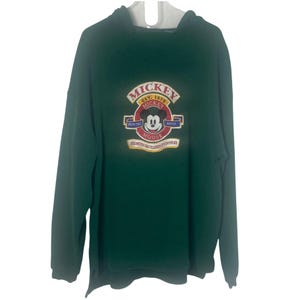 May include: A dark green long-sleeve hooded sweatshirt featuring a central embroidered graphic of Mickey Mouse. The design includes the text MICKEY EST. 1928, VERITAS ARTIS, and MICKEY MOUSE, with additional small text banners below the character.