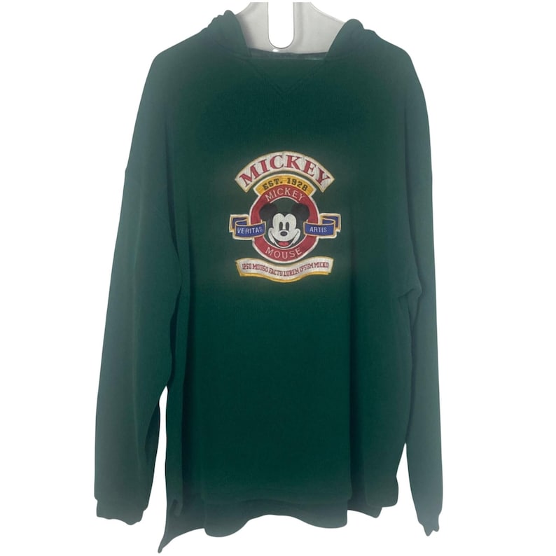 May include: A dark green long-sleeve hooded sweatshirt featuring a central embroidered graphic of Mickey Mouse. The design includes the text MICKEY EST. 1928, VERITAS ARTIS, and MICKEY MOUSE, with additional small text banners below the character.